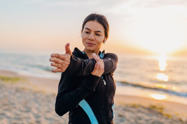 Joven y atractiva mujer delgada haciendo ejercicios deportivos en la playa del amanecer de la mañana en ropa deportiva, estilo de vida saludable, escuchando música en auriculares, haciendo estiramientos