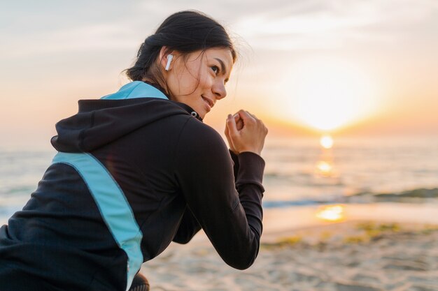 Joven y atractiva mujer delgada haciendo ejercicios deportivos en la playa del amanecer de la mañana en ropa deportiva, estilo de vida saludable, escuchando música con auriculares inalámbricos, haciendo abdominales