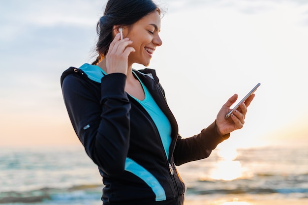 Joven y atractiva mujer delgada haciendo ejercicios deportivos en la playa del amanecer de la mañana en ropa deportiva, estilo de vida saludable, escuchando música en auriculares inalámbricos con smartphone, sonriendo feliz divirtiéndose