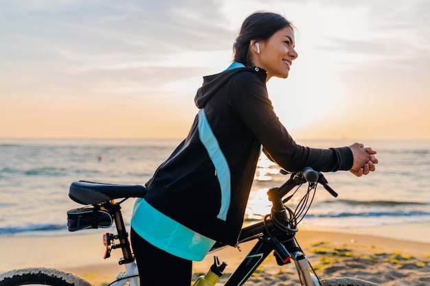 Joven y atractiva mujer delgada montando bicicleta, deporte en la playa de verano amanecer de mañana en ropa deportiva, estilo de vida activo y saludable, sonriendo feliz divirtiéndose