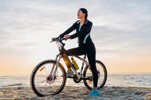 Foto gratis joven y atractiva mujer delgada montando bicicleta, deporte en la playa de verano amanecer de mañana en ropa deportiva, estilo de vida activo y saludable, sonriendo feliz divirtiéndose