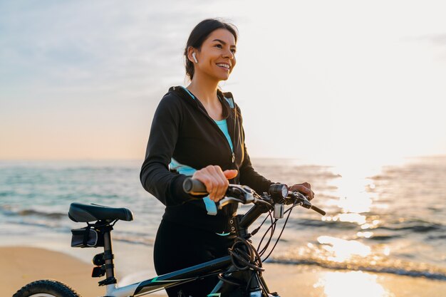 Joven y atractiva mujer delgada montando bicicleta, deporte en la playa de verano amanecer de mañana en ropa deportiva, estilo de vida activo y saludable, sonriendo feliz divirtiéndose