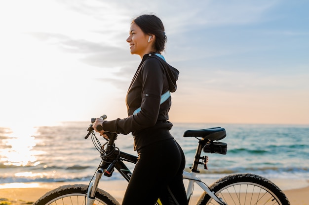 Joven y atractiva mujer delgada montando bicicleta, deportes en la playa del amanecer de la mañana en ropa deportiva deportiva, estilo de vida activo y saludable, sonriendo feliz divirtiéndose