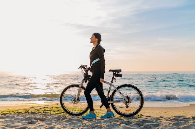 Joven y atractiva mujer delgada montando bicicleta, deportes en la playa del amanecer de la mañana en ropa deportiva deportiva, estilo de vida activo y saludable, sonriendo feliz divirtiéndose