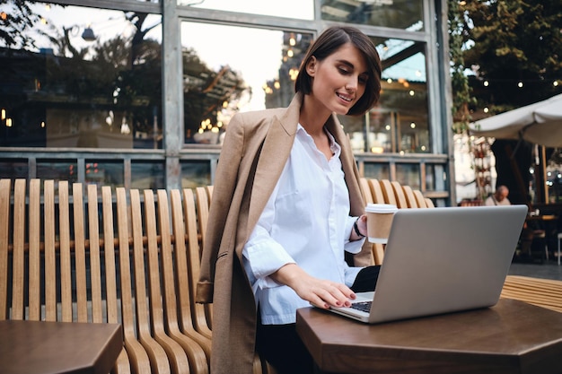 Joven y atractiva mujer de negocios sonriente felizmente trabajando en una laptop durante el descanso para tomar café en un café en la calle