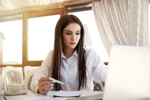 Foto gratuita joven atractiva mujer de pelo largo está trabajando en la computadora portátil con una taza de café