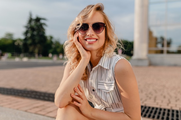 Joven y atractiva mujer rubia elegante linda sentada en las calles de la ciudad en vestido de algodón blanco de estilo de moda de verano con gafas de sol