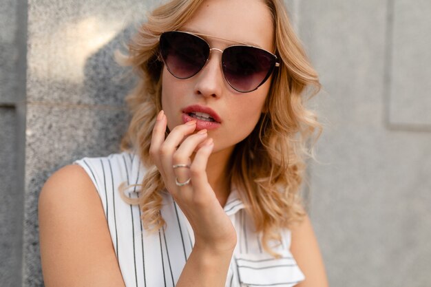 Joven y atractiva mujer rubia con estilo caminando en la calle de la ciudad en vestido de estilo de moda de verano con gafas de sol, mirada sexy