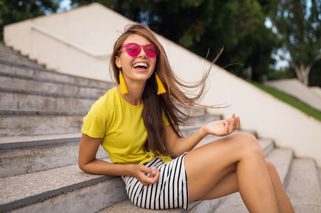 Joven y atractiva mujer sonriente elegante divirtiéndose en el parque de la ciudad, positivo, emocional, cabello largo, vistiendo top amarillo, minifalda a rayas, gafas de sol rosas, tendencia de moda de estilo veraniego,