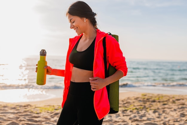 Joven y atractiva mujer sonriente haciendo deporte en el amanecer de la mañana en la playa del mar sosteniendo una estera de yoga y una botella de agua, estilo de vida saludable, escuchando música en auriculares, vistiendo una chaqueta cortavientos rosa