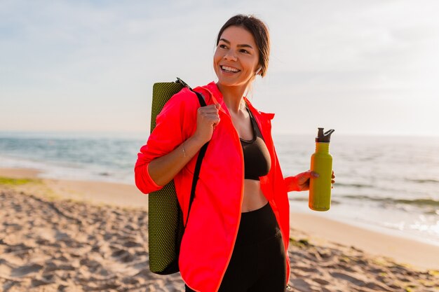 Joven y atractiva mujer sonriente haciendo deporte en el amanecer de la mañana en la playa del mar sosteniendo una estera de yoga y una botella de agua, estilo de vida saludable, escuchando música en auriculares, vistiendo una chaqueta cortavientos rosa