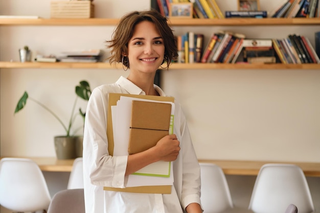 Foto gratuita joven atractiva mujer sonriente mirando alegremente a la cámara con papeles y bloc de notas en la oficina moderna