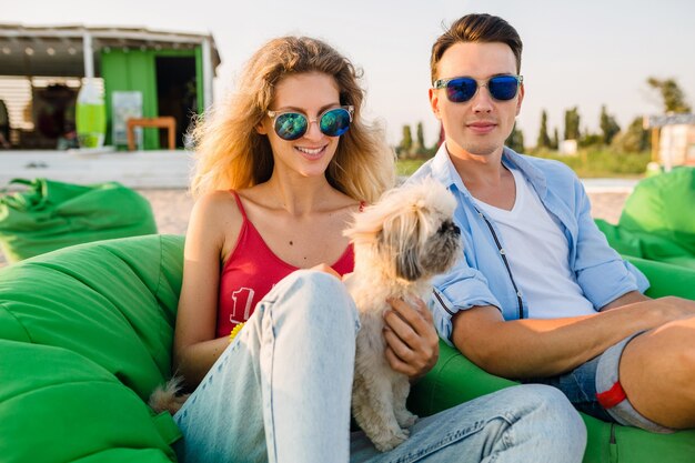 Joven y atractiva pareja sonriente divirtiéndose en la playa jugando con el perro de raza shih-tsu, sentado en una bolsa de frijoles verdes