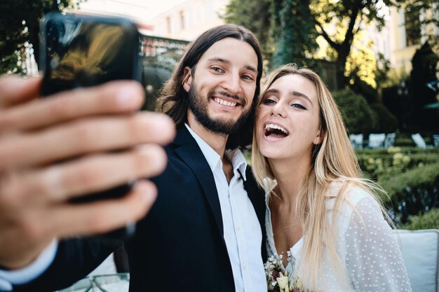 Joven atractivo alegre novio y novia felizmente tomando selfie juntos en el jardín del restaurante
