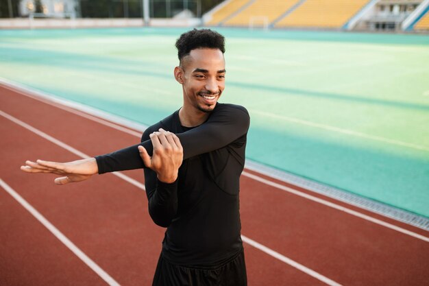 Joven y atractivo deportista afroamericano sonriente felizmente estirándose preparándose para correr en la pista de carreras en el estadio de la ciudad