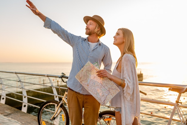 Joven atractivo y mujer viajando en bicicleta, sosteniendo mapa y turismo, pareja romántica en vacaciones de verano junto al mar al atardecer, amigos divirtiéndose juntos