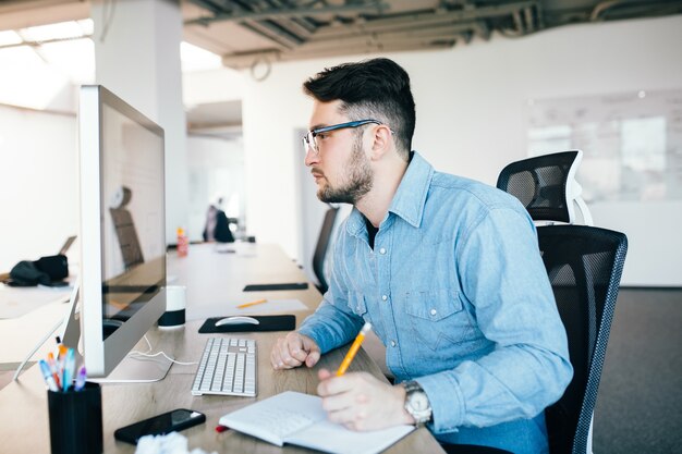 Joven atractivo de pelo oscuro en glassess está trabajando con una computadora en su lugar de trabajo en la oficina. Viste camisa azul. Parece ocupado, vista de lado.