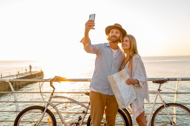 Joven atractivo sonriente feliz hombre y mujer viajando en bicicleta tomando foto selfie en la cámara del teléfono, pareja romántica junto al mar en la puesta de sol, traje de estilo boho hipster, amigos divirtiéndose juntos