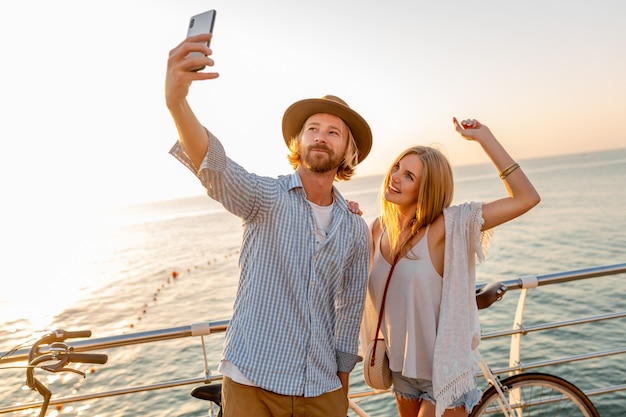 Joven atractivo sonriente feliz hombre y mujer viajando en bicicleta tomando fotos selfie en la cámara del teléfono, pareja romántica junto al mar en la puesta de sol, traje de estilo boho hipster, amigos divirtiéndose juntos