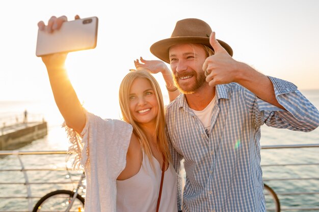 Joven atractivo sonriente feliz hombre y mujer viajando en bicicleta tomando fotos selfie en la cámara del teléfono, pareja romántica junto al mar en la puesta de sol, traje de estilo boho hipster, amigos divirtiéndose juntos