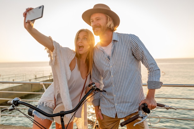 Joven atractivo sonriente feliz hombre y mujer viajando en bicicleta tomando fotos selfie en la cámara del teléfono, pareja romántica junto al mar en la puesta de sol, traje de estilo boho hipster, amigos divirtiéndose juntos