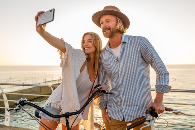 Joven atractivo sonriente feliz hombre y mujer viajando en bicicleta tomando fotos selfie en la cámara del teléfono, pareja romántica junto al mar en la puesta de sol, traje de estilo boho hipster, amigos divirtiéndose juntos