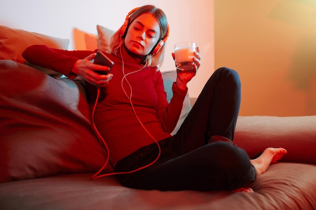 Joven con auriculares sentada en el sofá con un vaso de leche y un celular en las manos Una chica bonita escuchando música mientras pasa tiempo en casa