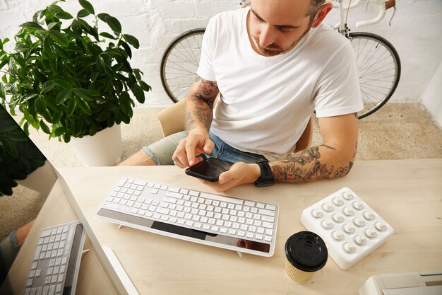 Joven autónomo tatuado en camiseta blanca en blanco usa su teléfono móvil cerca de su computadora en casa frente a una pared de ladrillo y una bicicleta vintage estacionada, horario de verano