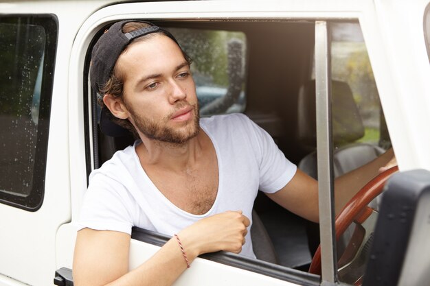 Joven aventurero barbudo con snapback negro conduciendo su SUV blanco, yendo a un viaje de safari, asomando la cabeza por la ventana abierta, luciendo cansado e infeliz