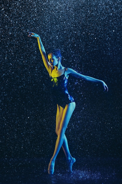 Joven bailarina de ballet actuando bajo gotas de agua y spray. Modelo caucásico bailando en luces de neón. Mujer atractiva. Concepto de ballet y coreografía contemporánea.