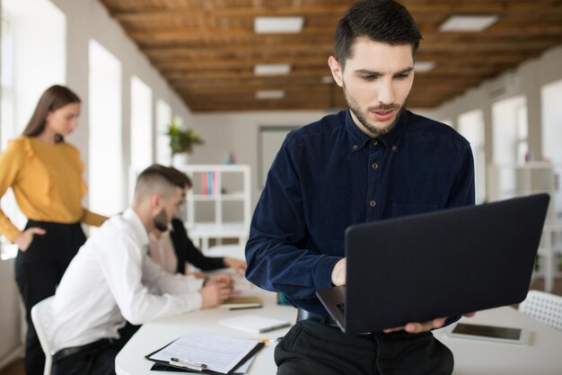 Joven con barba en camisa cuidadosamente usando una computadora portátil mientras pasa tiempo en la oficina con colegas en el fondo