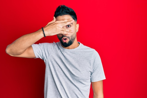 Foto gratuita joven con barba con camiseta blanca informal mirando con sorpresa cubriendo la cara y los ojos con la mano, mirando a través de los dedos con miedo