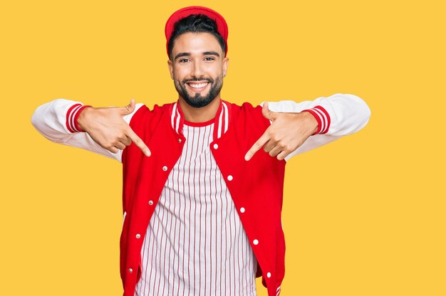 Joven con barba con uniforme de béisbol que se ve confiado con una sonrisa en la cara, apuntándose con los dedos orgulloso y feliz.