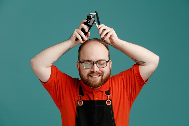 Foto gratuita joven barbero complacido con gafas, camisa roja y delantal de barbero sosteniendo herramientas de barbería en la cabeza sonriendo mientras mantiene los ojos cerrados aislados en el fondo azul