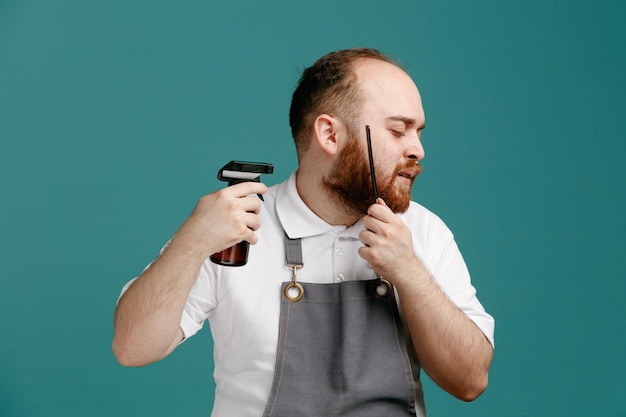 Joven barbero concentrado con camisa blanca y delantal de barbero girando la cabeza hacia un lado peinándose la barba y aplicando spray para el cabello en su propia barba con los ojos cerrados aislados en el fondo azul