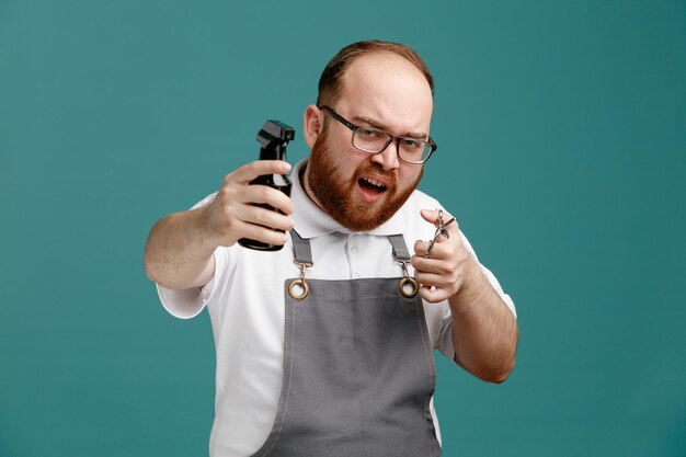 Joven barbero frunciendo el ceño con uniforme y gafas sosteniendo tijeras mirando a la cámara estirando la botella de spray hacia la cámara aislada en el fondo azul