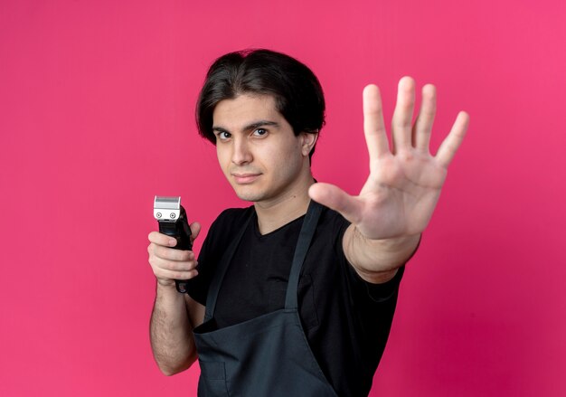 Joven barbero guapo en uniforme sosteniendo cortapelos y extendiendo la mano aislada en la pared rosa