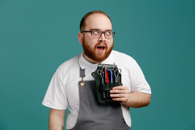 Joven barbero impresionado con uniforme y gafas sosteniendo una bolsa de barbero con herramientas de barbería mirando al lado aislado en el fondo azul