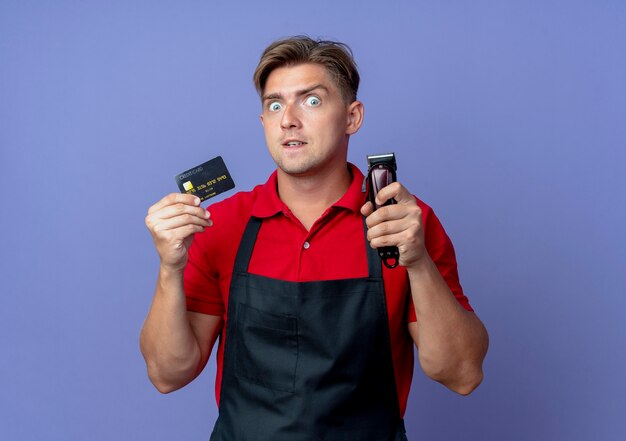 Joven barbero macho rubio sorprendido en uniforme tiene tarjeta de crédito y cortapelos aislado