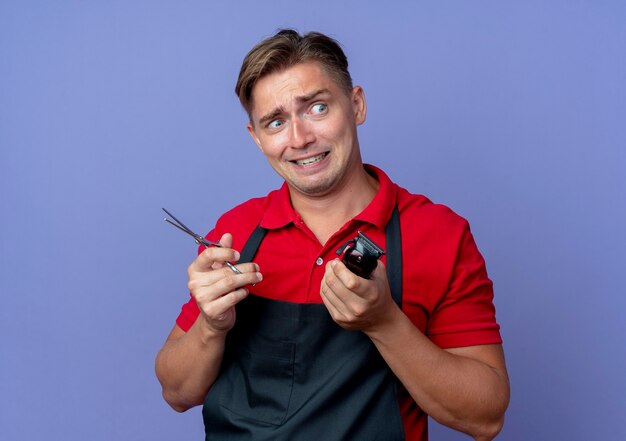 Foto gratuita joven barbero masculino rubio asustado en uniforme sostiene tijeras y cortadora de cabello mirando al lado aislado en el espacio violeta con espacio de copia