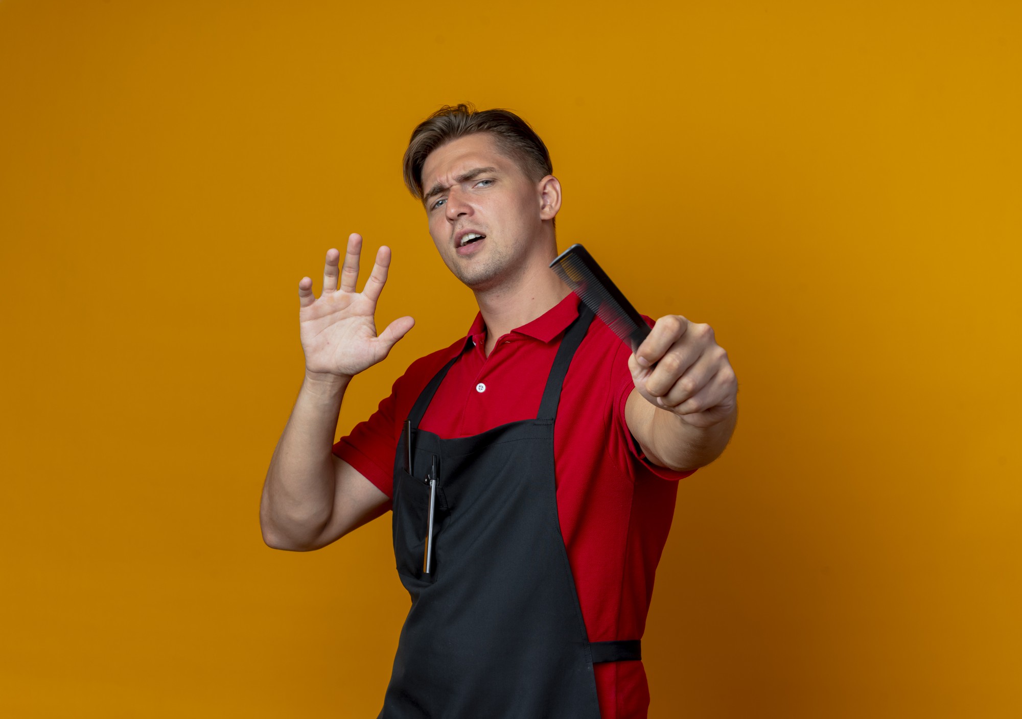 Joven barbero masculino rubio molesto en uniforme sostiene el peine estirando las manos aisladas sobre fondo naranja con espacio de copia