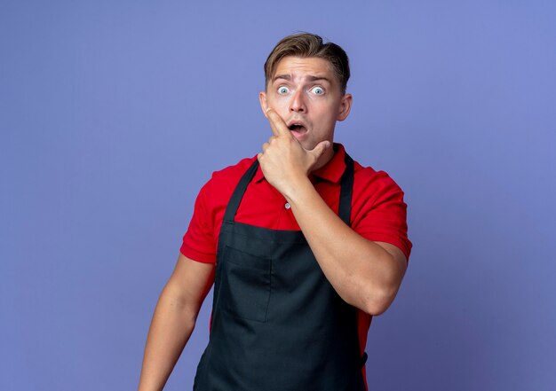 Joven barbero masculino rubio sorprendido en uniforme pone la mano en la barbilla aislada en el espacio violeta con espacio de copia