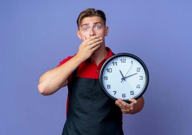 Joven barbero masculino rubio sorprendido en uniforme pone la mano en la boca sosteniendo el reloj aislado en el espacio violeta con espacio de copia