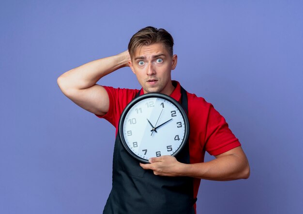 Joven barbero masculino rubio sorprendido en uniforme pone la mano detrás de la cabeza sosteniendo el reloj aislado en el espacio violeta con espacio de copia