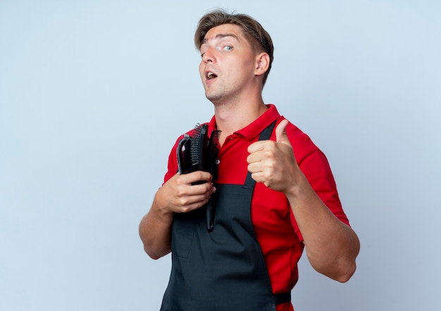 Joven barbero masculino rubio sorprendido en uniforme pulgar hacia arriba sosteniendo herramientas de peluquero aisladas en espacio en blanco con espacio de copia