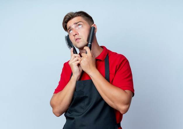 Joven barbero masculino rubio en uniforme pone peinetas en la cara mirando hacia arriba