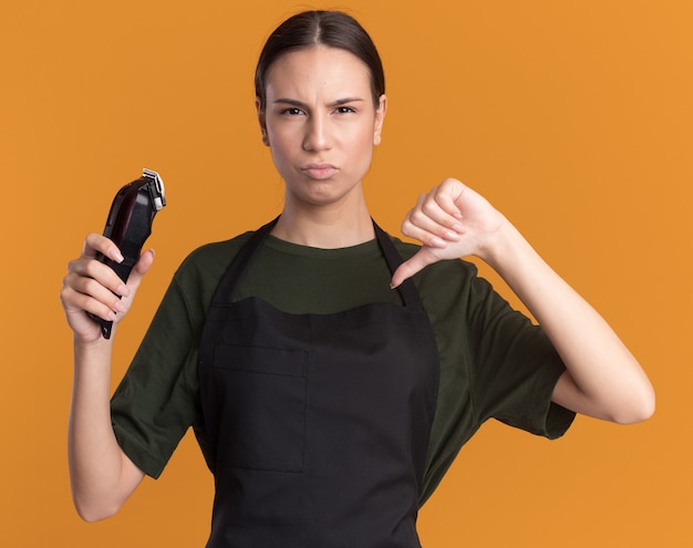 Joven barbero morena disgustada en uniforme con los pulgares hacia abajo y sostiene cortapelos aislado en la pared naranja con espacio de copia