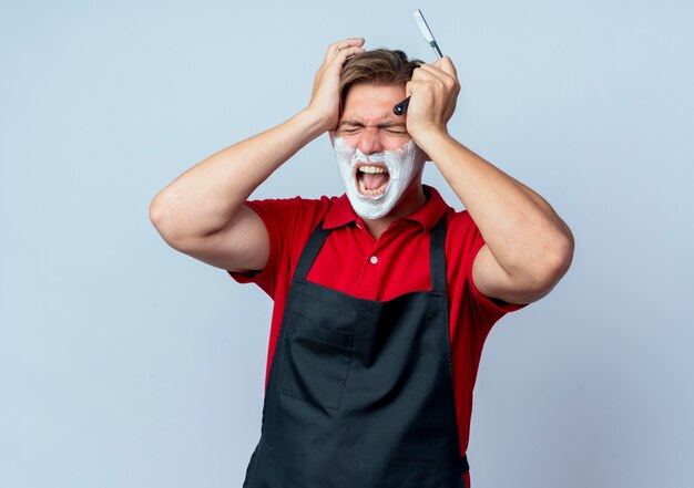 Joven barbero rubio molesto en uniforme con la cara manchada con espuma de afeitar sostiene la navaja de afeitar pone las manos en la cabeza aislada en el espacio en blanco con espacio de copia