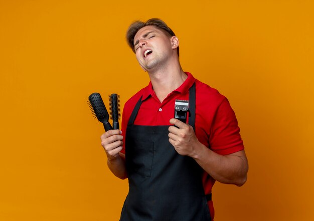 Joven barbero rubio molesto en uniforme sostiene peines y cortadora de cabello aislado en espacio naranja con espacio de copia