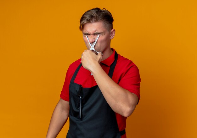 Joven barbero rubio molesto en uniforme sostiene tijeras en la nariz pretendiendo cortar aislado en espacio naranja con espacio de copia